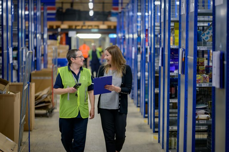Two workers walk through a warehouse, discussing details while checking documents.
