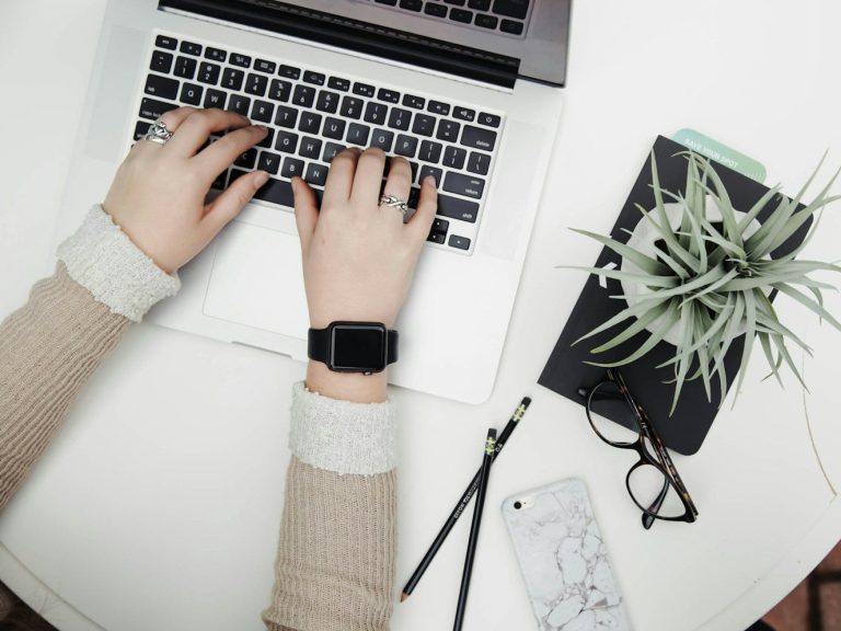 Person typing on a laptop with a notebook and plant nearby on a desk.