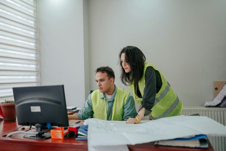 Two workers in high-visibility vests reviewing plans at a computer desk.