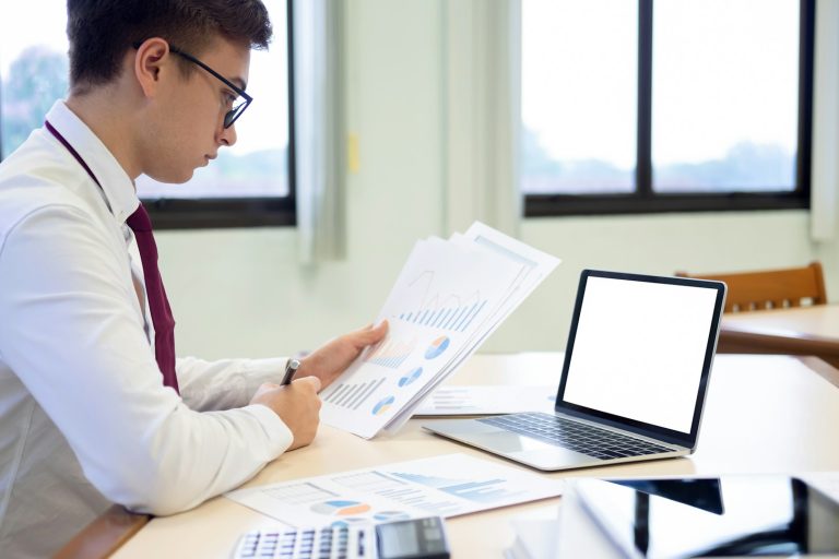 A man in a white shirt and tie studies documents next to an empty laptop on a table.