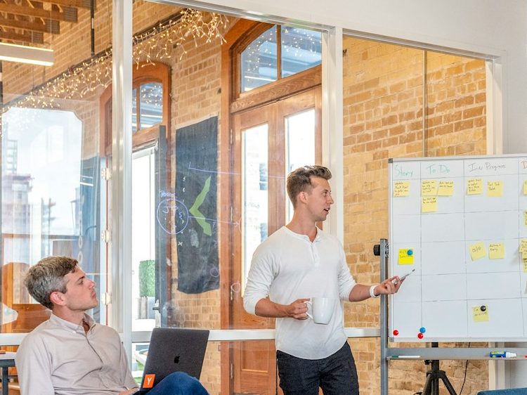 A man presents ideas on a whiteboard while another listens, in a modern office setting.